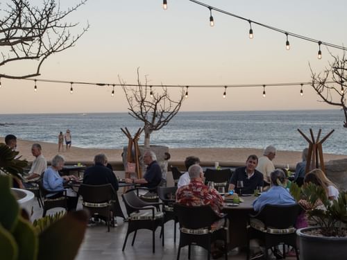 Comensales cenando en un patio bajo luces de cadena con vista al mar en De Cortez Grill House en Hacienda Del Mar Los Cabos.