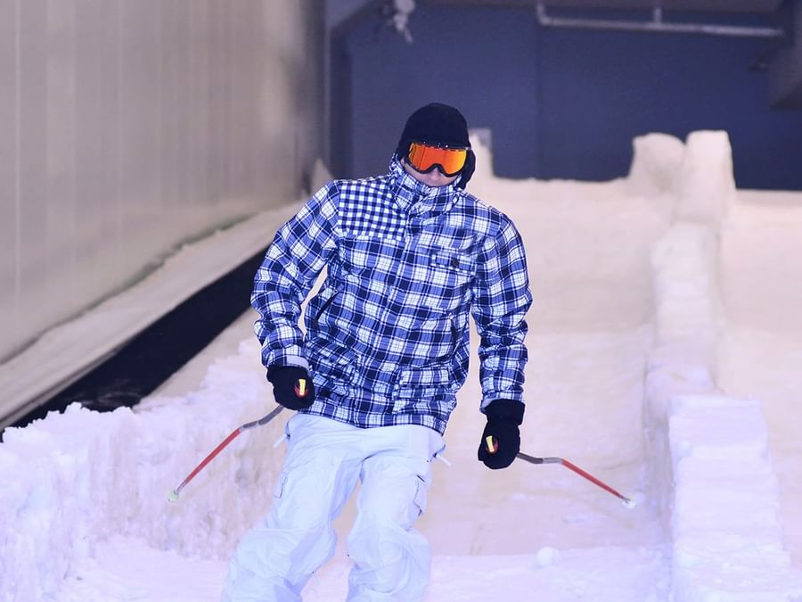 Person in a plaid jacket and white pants skiing on an indoor snow track near DAMAC Hotels & Resorts