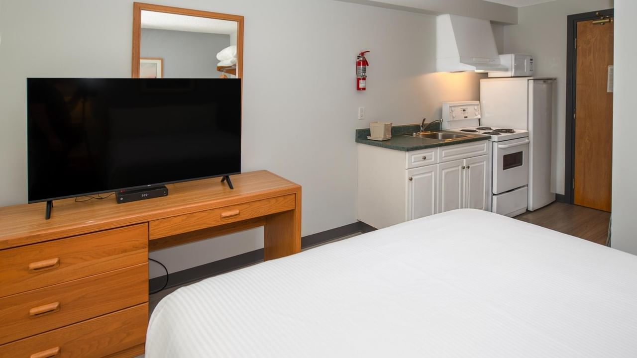 Room with white bed, flat-screen TV on wooden desk, kitchen area with sink and stove.
