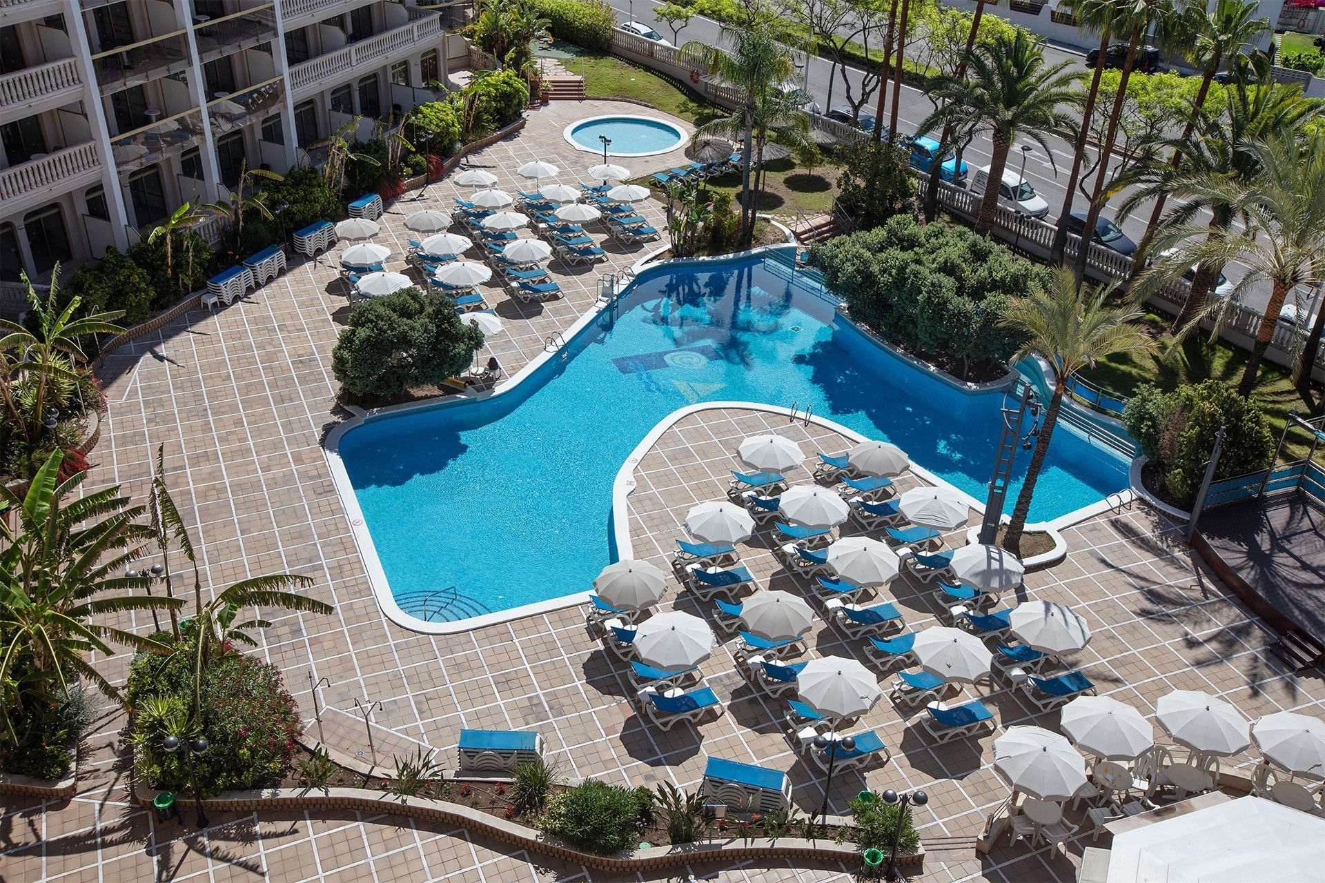 High-angle view of a bright blue, resort-style swimming pool surrounded by dozens of white sun umbrellas and lounge chairs.