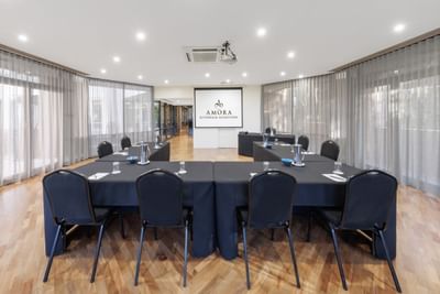 Professional meeting room, with a U-shaped table setup and a branded presentation screen at Amora Hotel Group