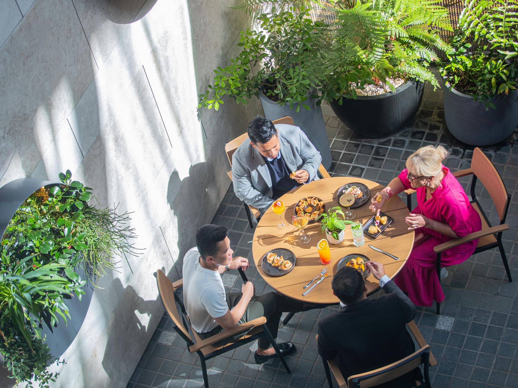 Four people sit around a table in an outdoor dining area with potted plants and natural light.