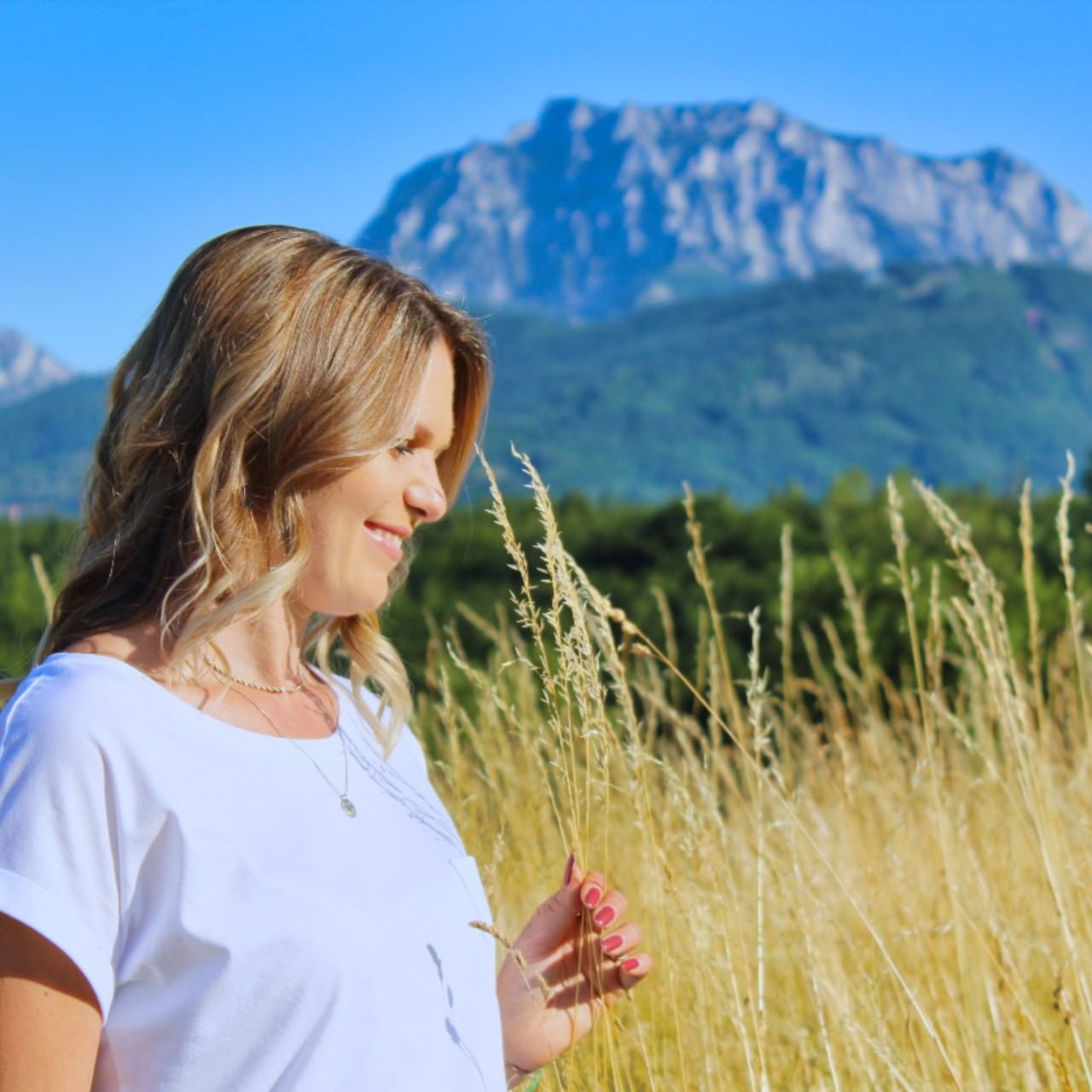 Lächelnde Frau in einem Feld mit Bergen im Hintergrund, sie hält eine Grashalm.