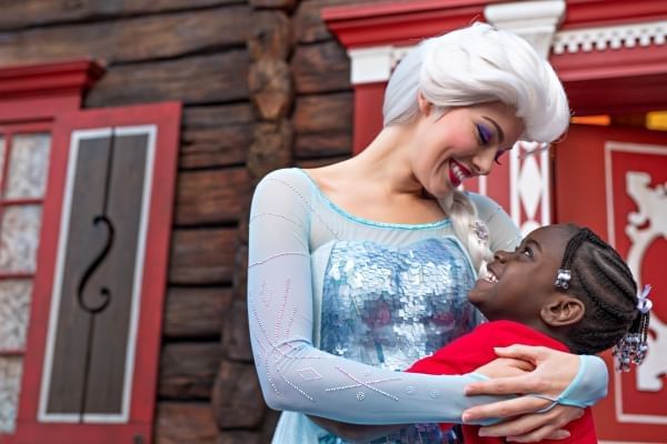 Elsa, in a beautiful light blue sparkly dress, smiles and embraces a smiling girl in front of a rustic brown building with red and white detailing. 