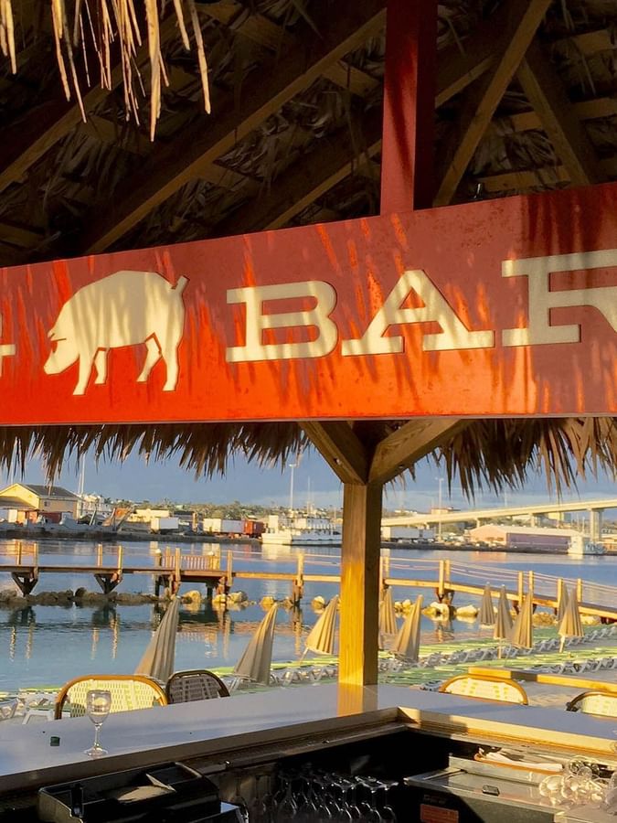 Hog Bar sign under a thatched roof by a calm bay and wooden pie at Warwick Paradise Island Bahamas