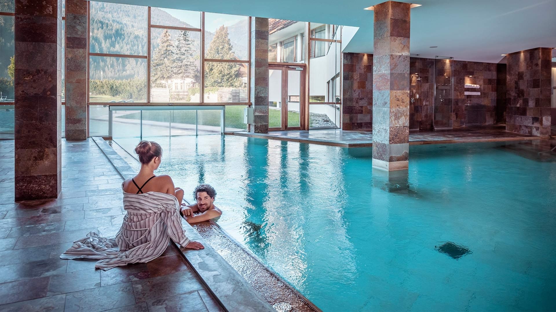 Indoor pool with a woman sitting and a man swimming, surrounded by stone columns and large windows.