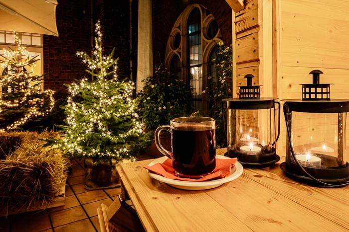 Tasse mit Glühwein auf Holztisch vor Weihnachtsbäumen mit Lichterketten und Kerzen.