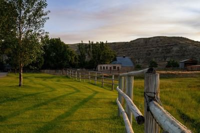 Wooden fence line in a verdant field leading to buildings with hills in the background at The Ranch at Ucross