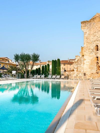 An outdoor pool flanked by lounge chairs and historic stone walls with buildings in background.