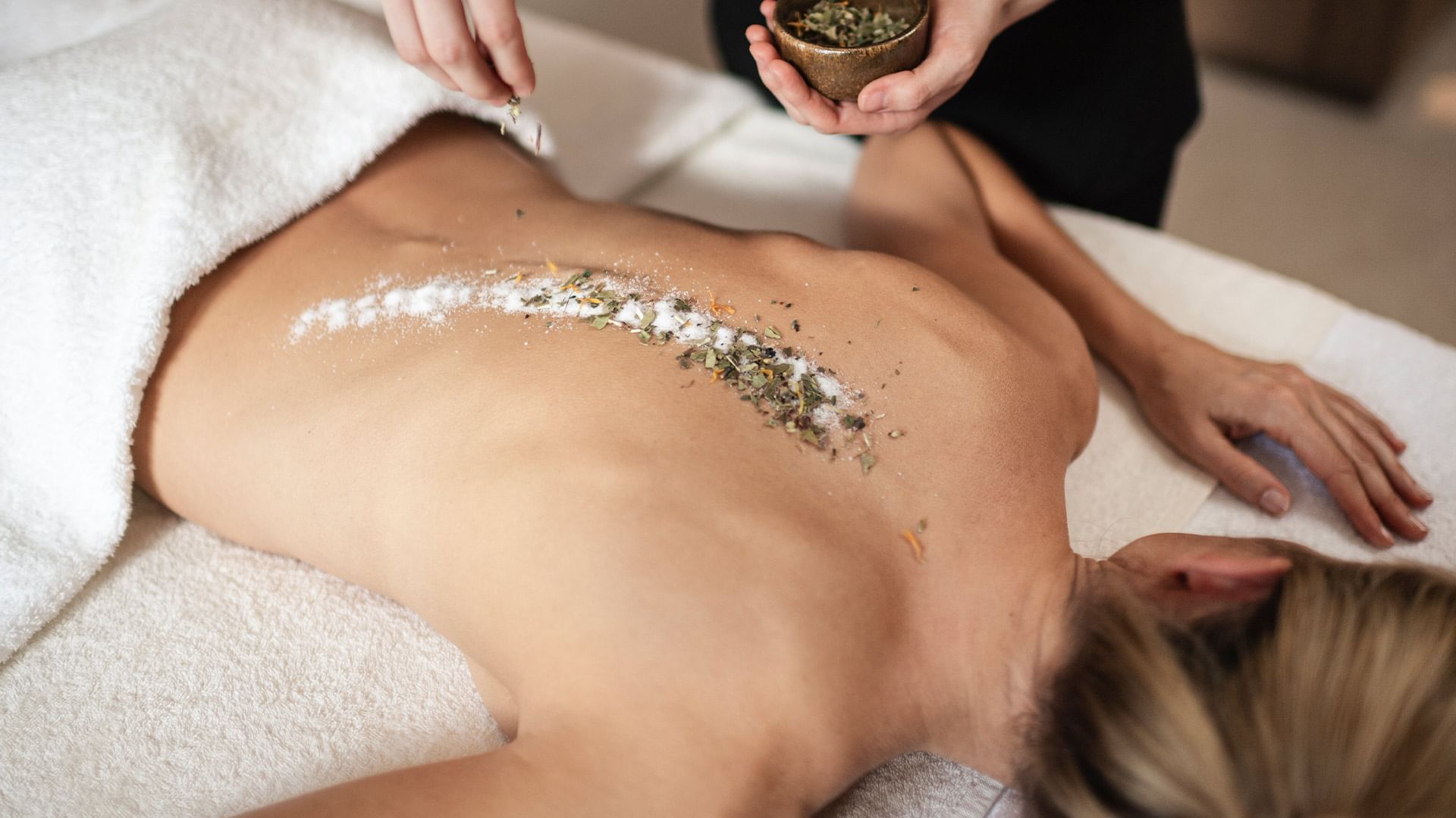 Frau liegt auf dem Rücken während einer Massage, jemand streut Kräuter auf ihren Rücken.