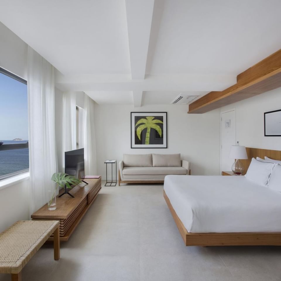 suíte master casal com vista para o mar do Janeiro Hotel decorada com uma bela cama king size com um lindo quadro do Cristo Redentor, sofá em tons claros e TV  