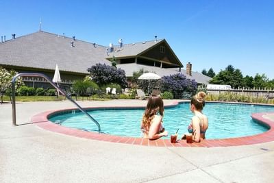 Two women in bikinis sit poolside with drinks at an outdoor pool with oregon garden resort in the background.