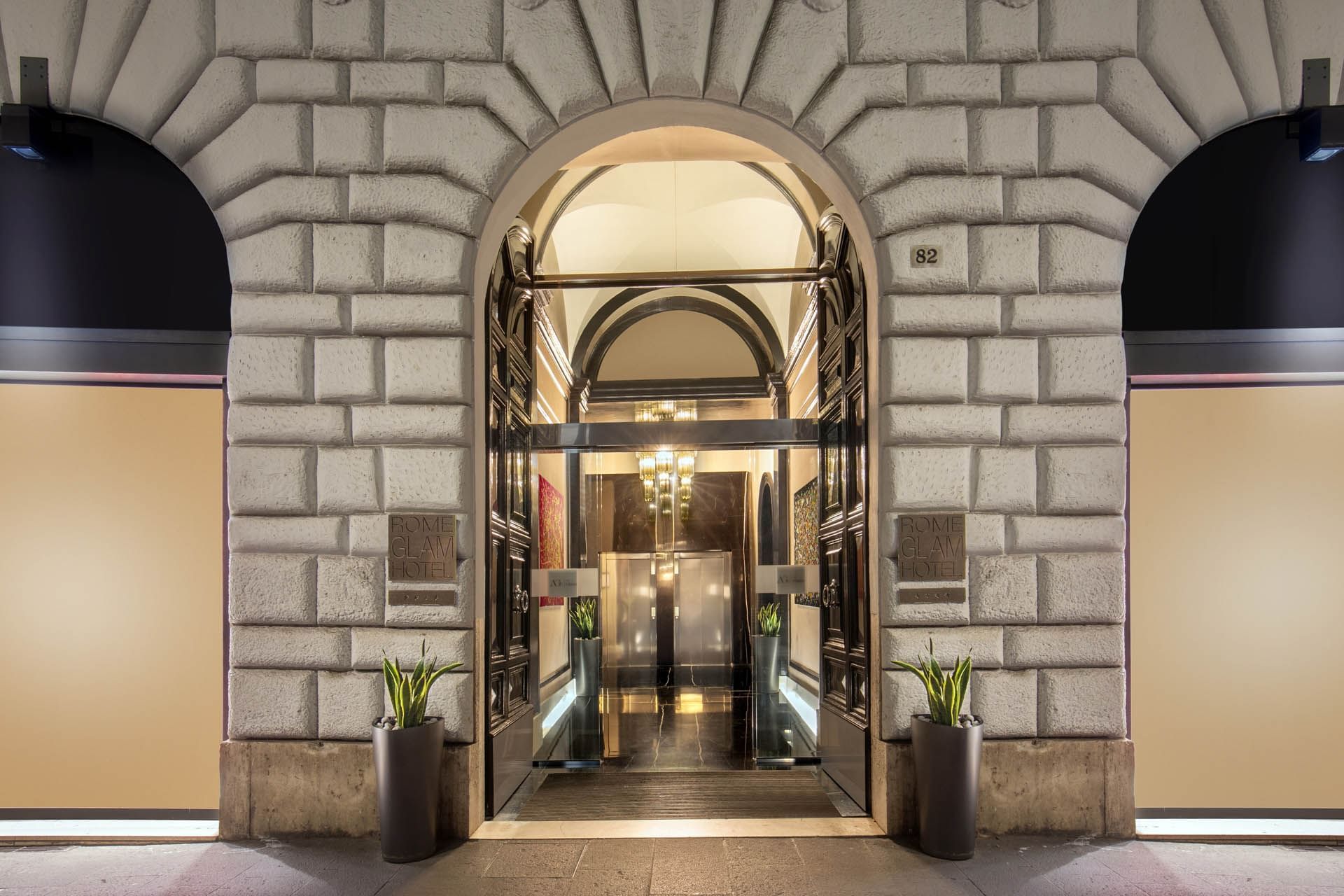 Elegant entrance of The Glam Hotel, featuring stone architecture, glass doors, and decorative planters
