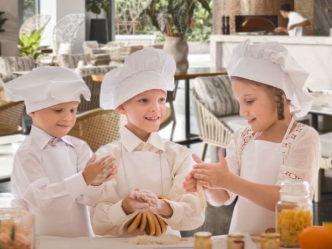 Three children in chef hats and aprons happily kneading dough on a kitchen counter.