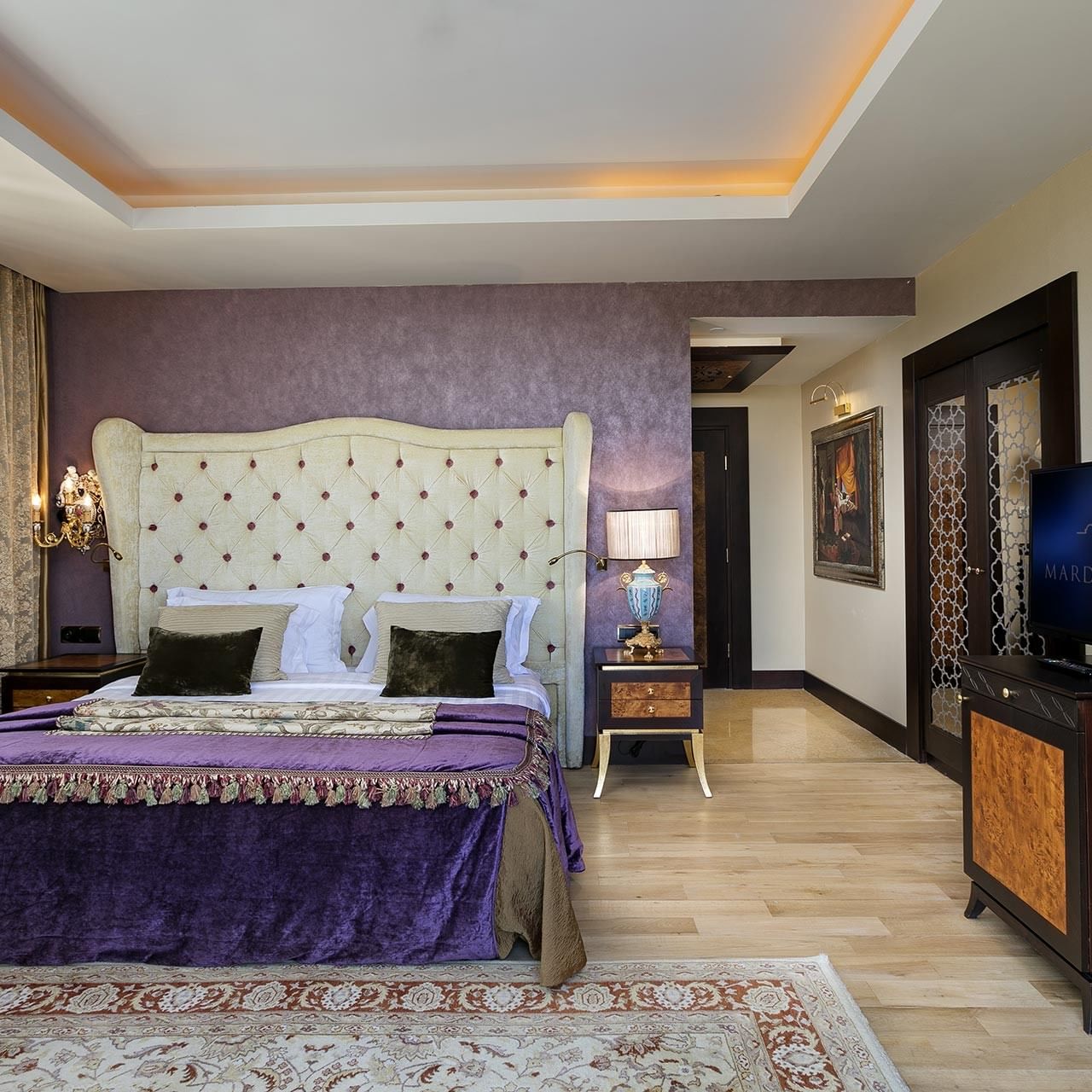 Роскошная спальня с большой кроватью у фиолетовой стены, декоративным изголовьем, телевизором и ковром