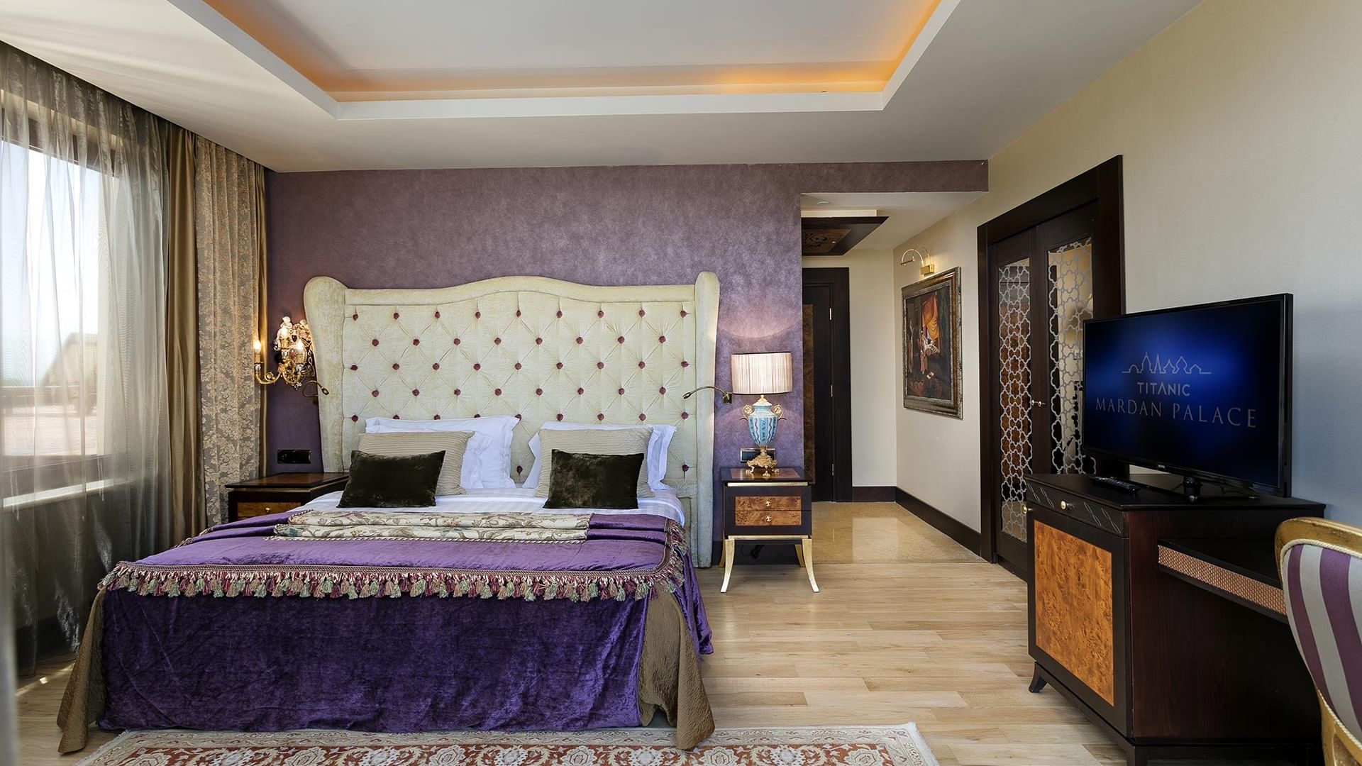 Роскошная спальня с большой кроватью у фиолетовой стены, декоративным изголовьем, телевизором и ковром