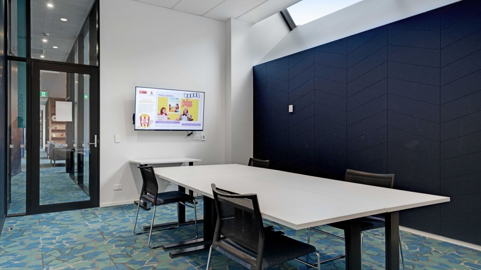 Modern meeting room with white table, black chairs, wall-mounted screen, and blue accent wall at UniLodge Auckland Central.