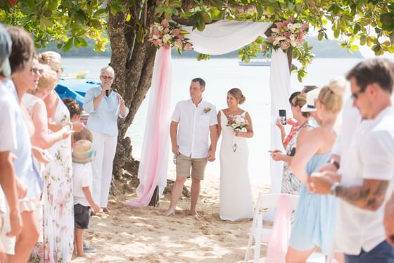 Wedded couple standing hand in hand under a floral arch on the beach, surrounded by guests at Bay Gardens Hotels and Resorts
