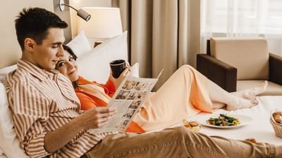 Couple relaxing on bed with a newspaper, coffee, and a plate of food at Polonia Palace Hotel