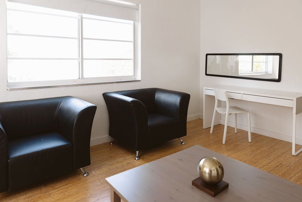 A bright living room with two black leather chairs, a wooden coffee table, and a white desk with a mirror above it.