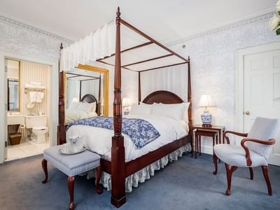 Four-poster bed with blue quilt by a striped bench and armchair near mirror in Deluxe Queen at Simsbury 1820 House