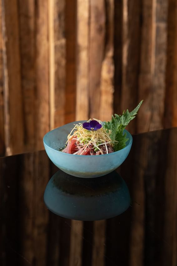 Sashimi and sprouts bowl with a purple flower on a black surface at The Kitchens