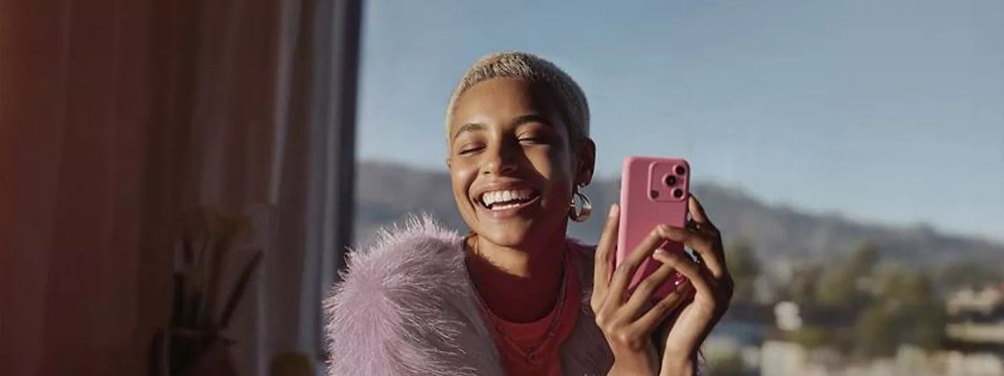 Woman smiling and taking a selfie with a pink smartphone near a window.