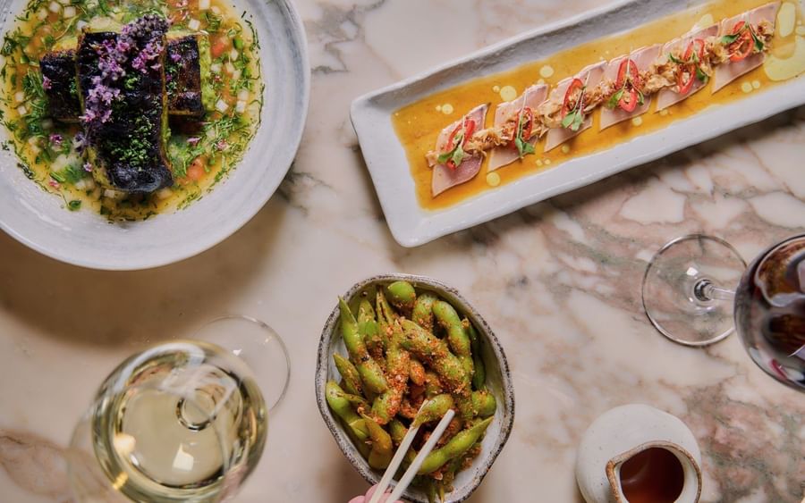 Overhead view of gourmet dishes including seared fish, cured meat with chili, and edamame on marble table at May Fair Kitchen
