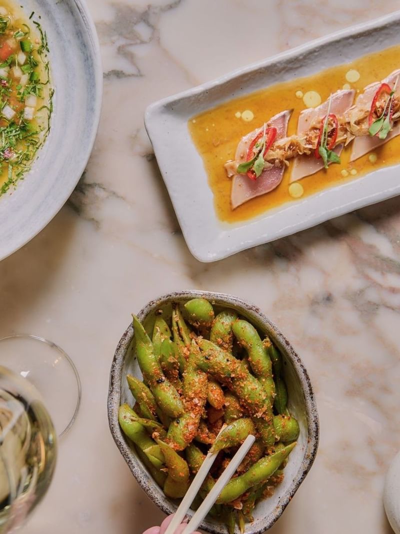 Bowl of edamame with chopsticks, two plates of food, and two wine glasses on a table at May Fair Kitchen