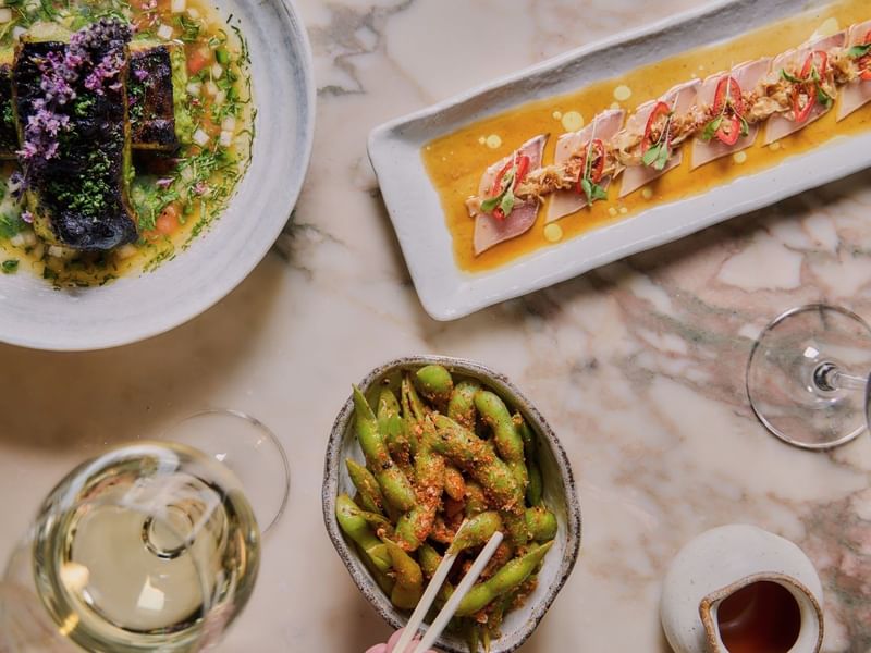 Bowl of edamame with chopsticks, two plates of food, and two wine glasses on a table at May Fair Kitchen
