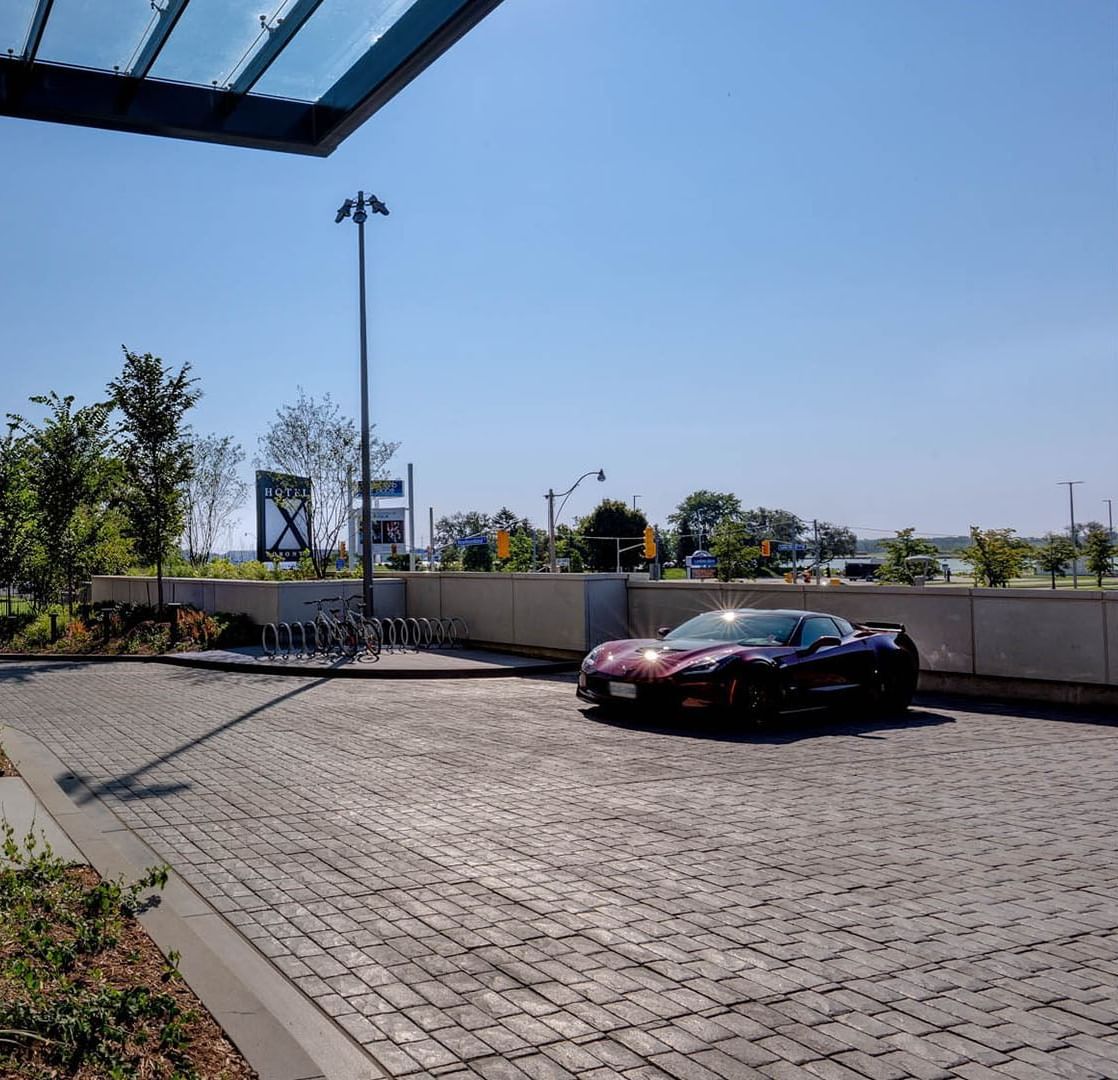 Sleek dark sports car parked on stone driveway facing entrance canopy and city skyline views at Hotel X Toronto