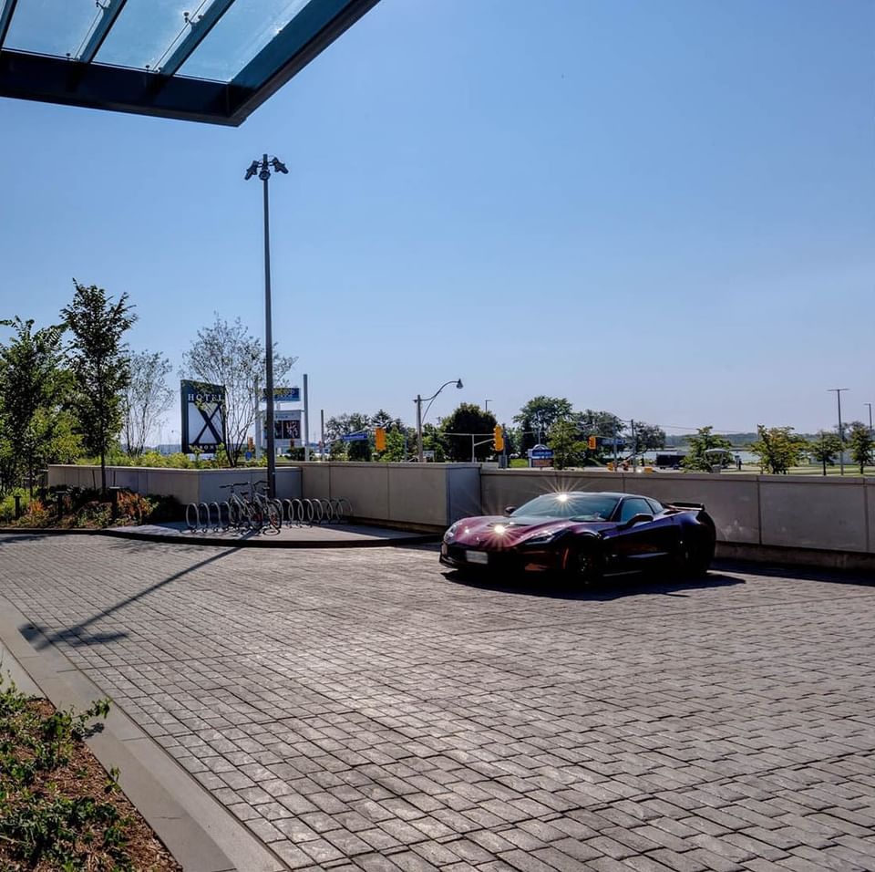 Sleek dark sports car parked on stone driveway facing entrance canopy and city skyline views at Hotel X Toronto