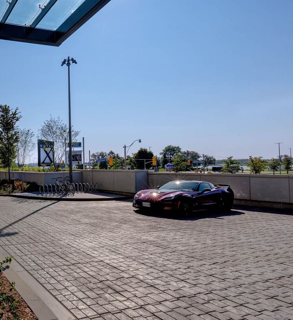 Sleek dark sports car parked on stone driveway facing entrance canopy and city skyline views at Hotel X Toronto