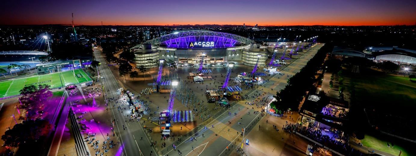 Aerial view of Accor Stadium at twilight, with vibrant purple lights, crowds, and event setups near Novotel Sydney Olympic Park.