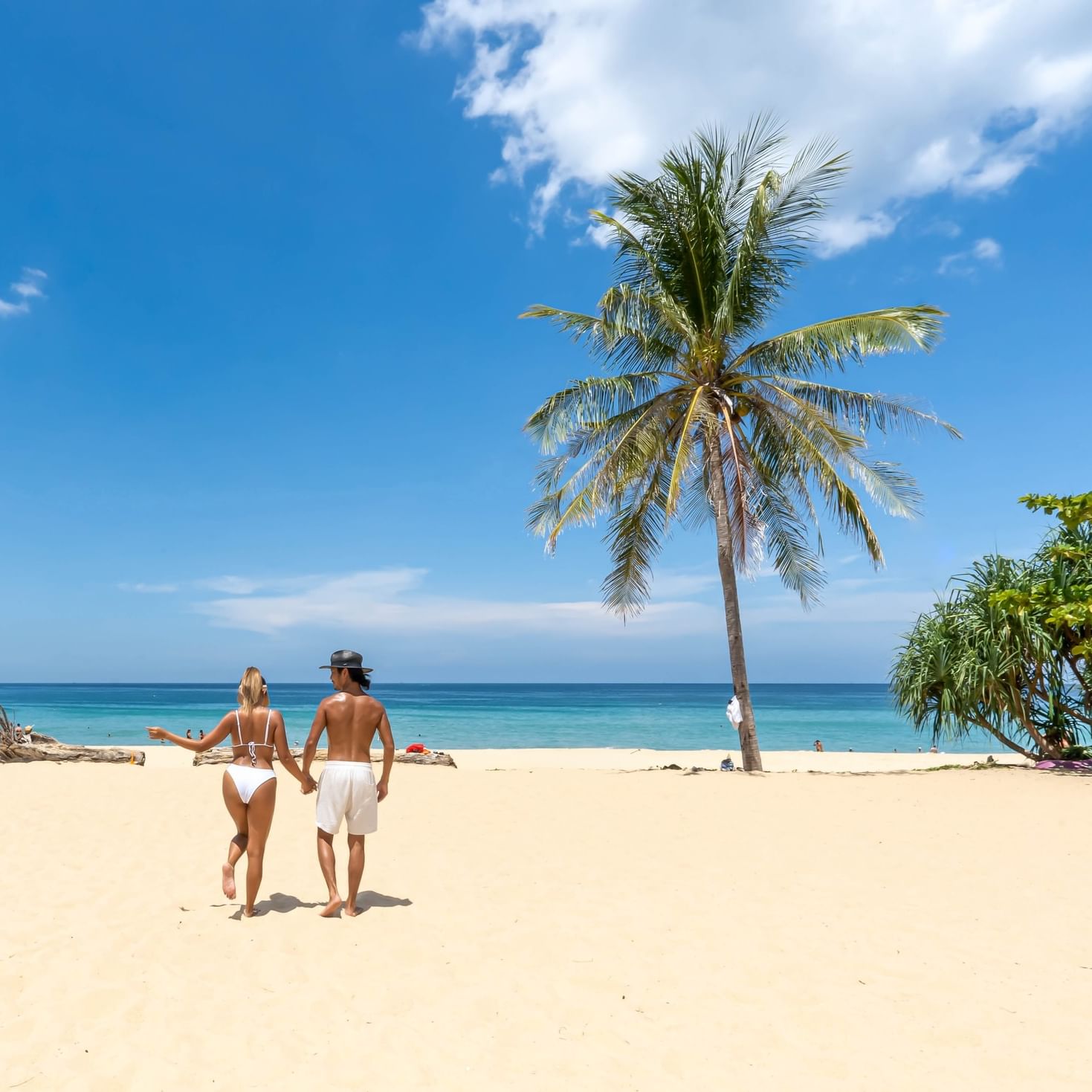 Couple walking hand-in-hand on the soft sand of Karon Beach Phuket, with clear blue skies, turquoise sea, and a palm tree creating a perfect tropical getaway scene.