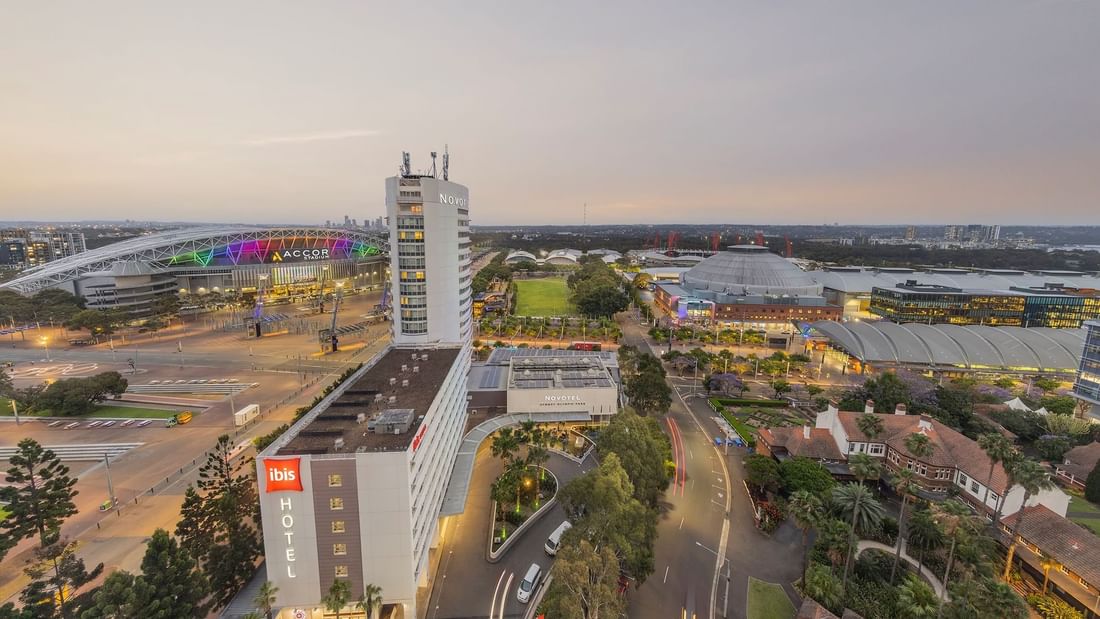 Aerial view of Novotel Sydney Olympic Park and Accor Stadium featuring vibrant lights, lush greenery, and city skyline