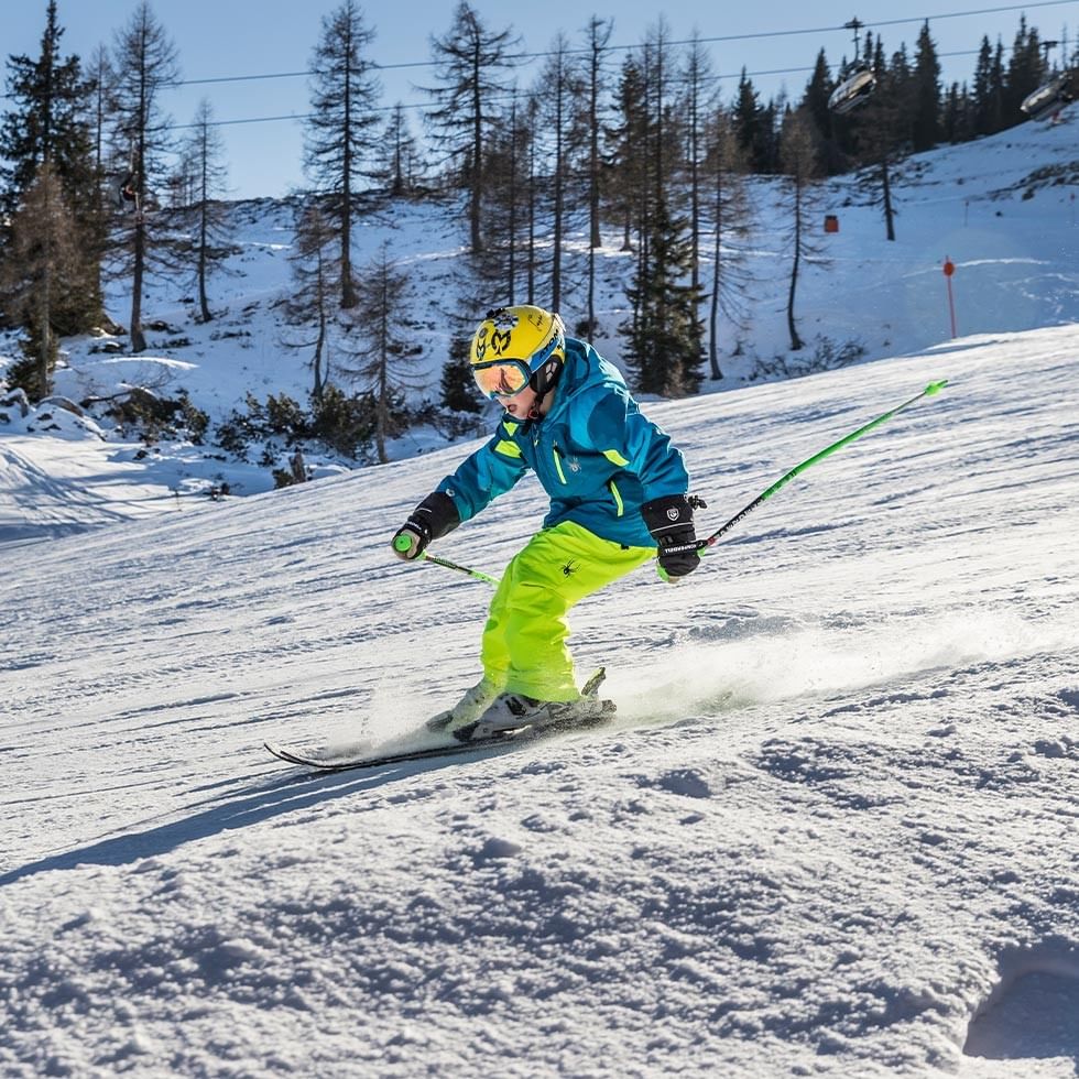 Skier in blue jacket and yellow pants skiing down snowy slope with trees in the background.
