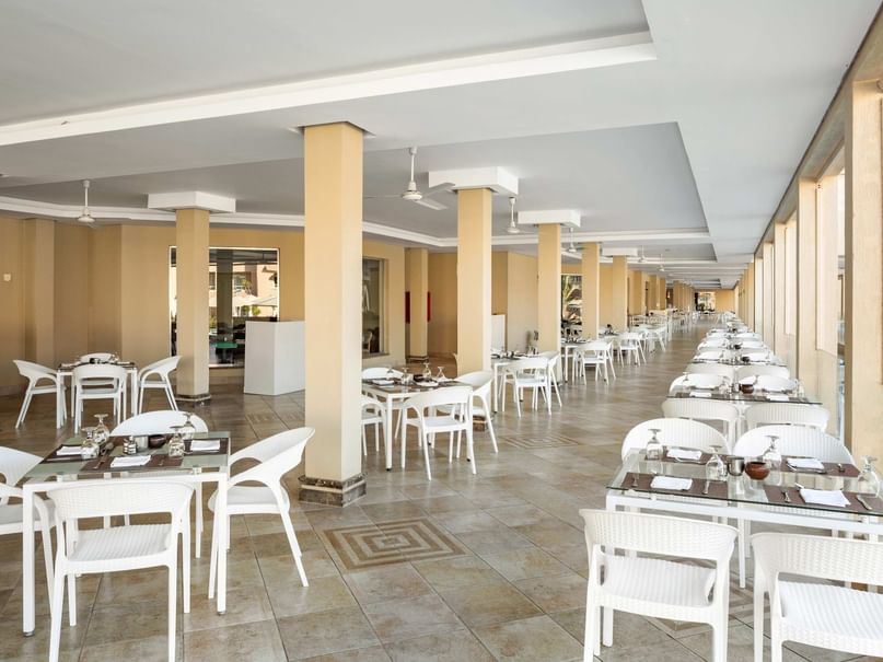 Leeres Restaurant mit weißen Tischen und Stühlen, neben einem Pool gelegen, mit hellen Fliesen und Säulen.