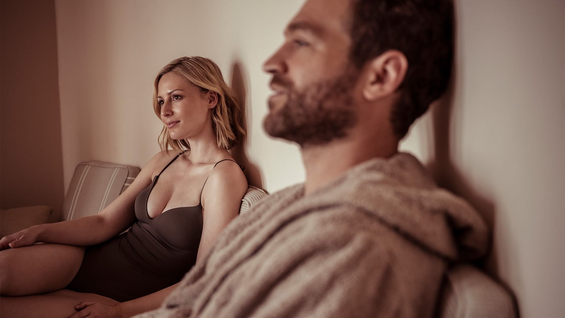 Mann und Frau sitzen auf Couch, schauen sich an, Mann trägt Bademantel, Frau trägt Kleid.