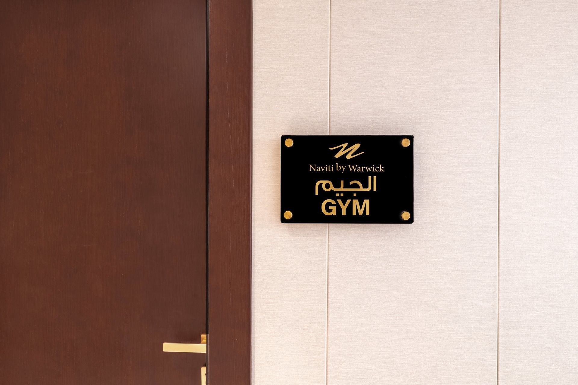 Black acrylic Gym door sign plaque with Arabic script mounted on wall beside wooden entryway at Naviti by Warwick Jeddah