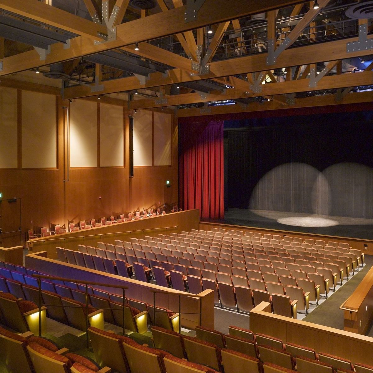An empty theater with wooden chairs and a stage with red curtains at the back.