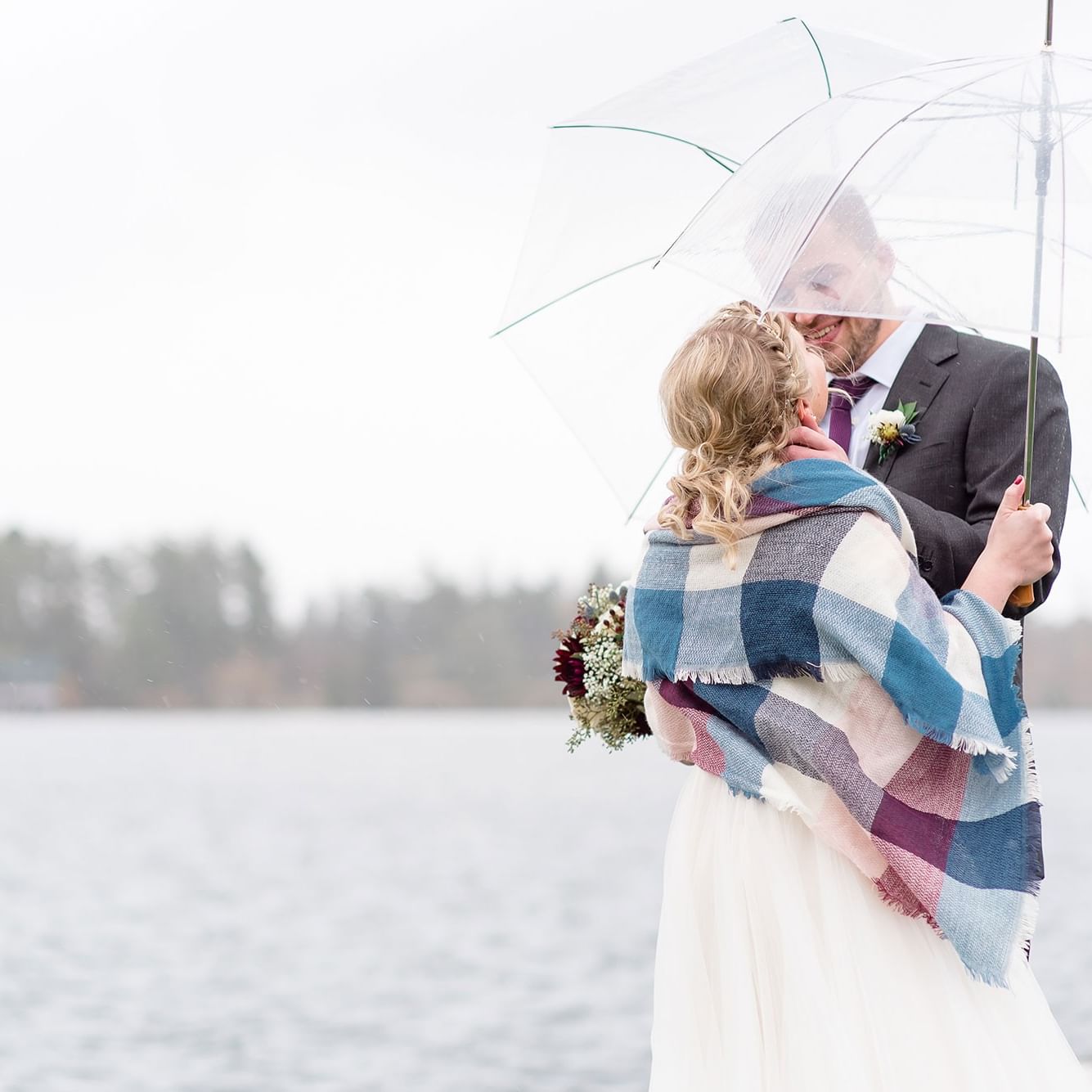 Bridal couple under umbrella, bride with bouquet and plaid blanket, by lake with trees in background.