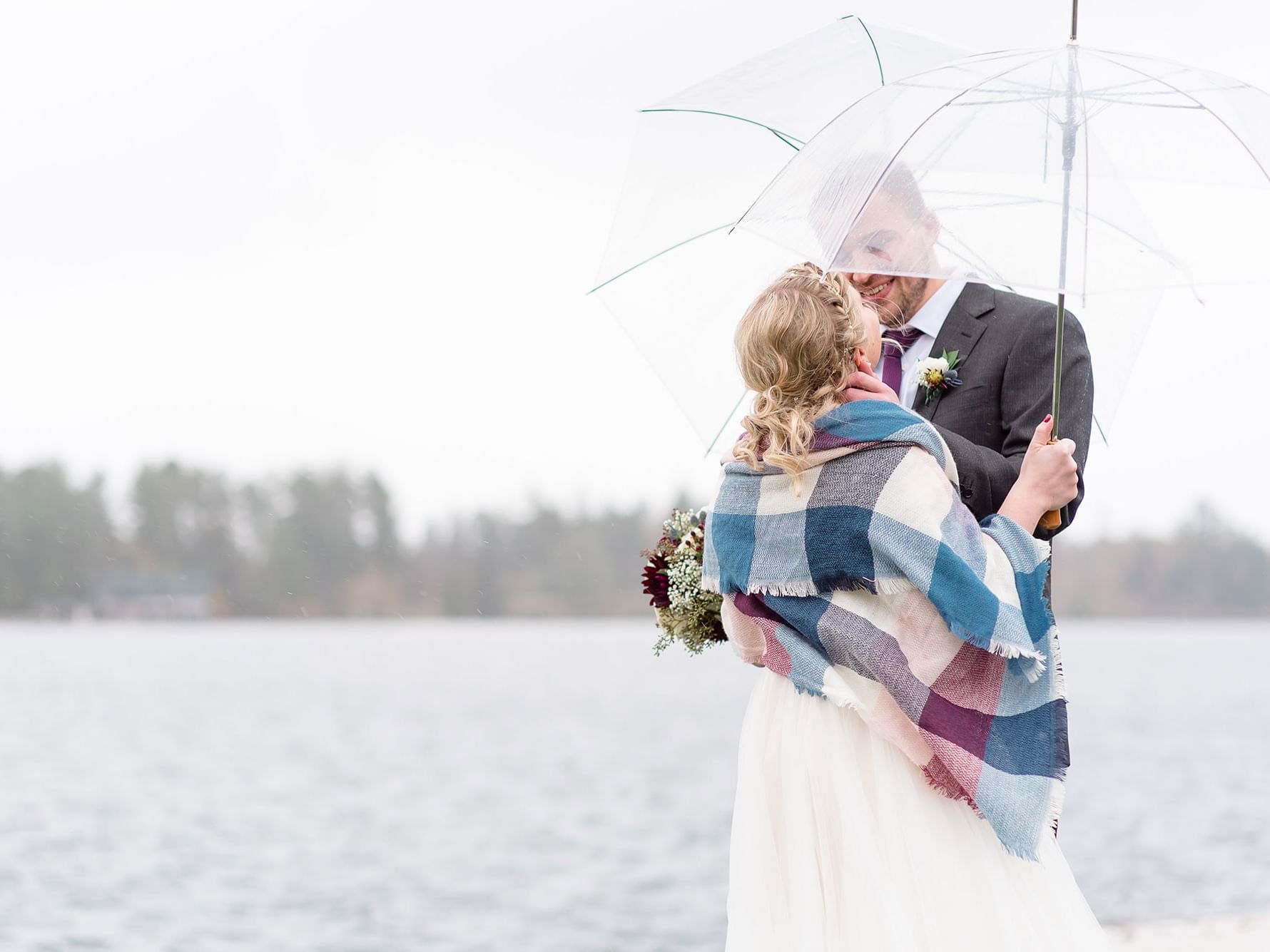 Bridal couple under umbrella, bride with bouquet and plaid blanket, by lake with trees in background.