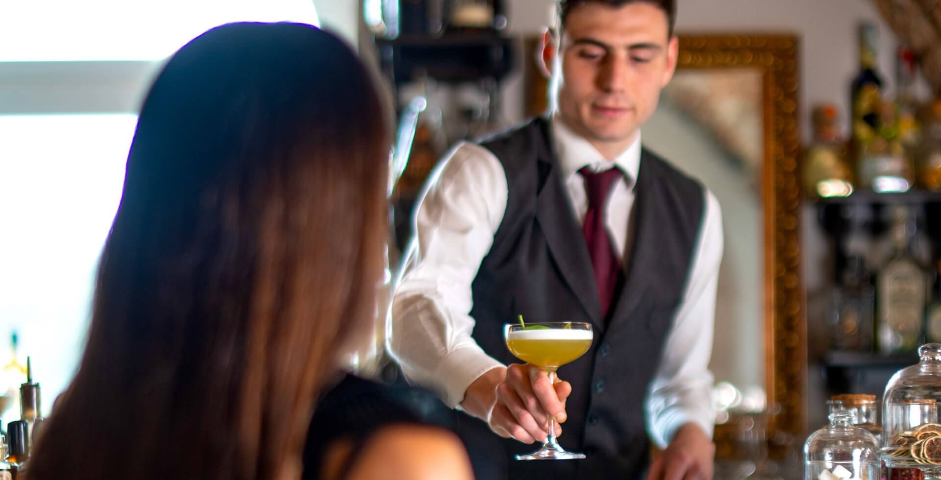 Bartender in vest and tie serves a cocktail to a woman in a bar.