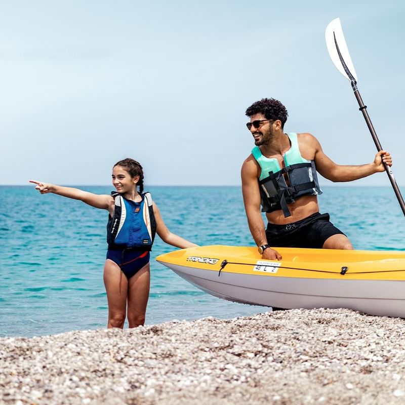 Uomo e bambina con giubbotto di salvataggio e kayak sulla spiaggia, pronti per un'avventura acquatica.