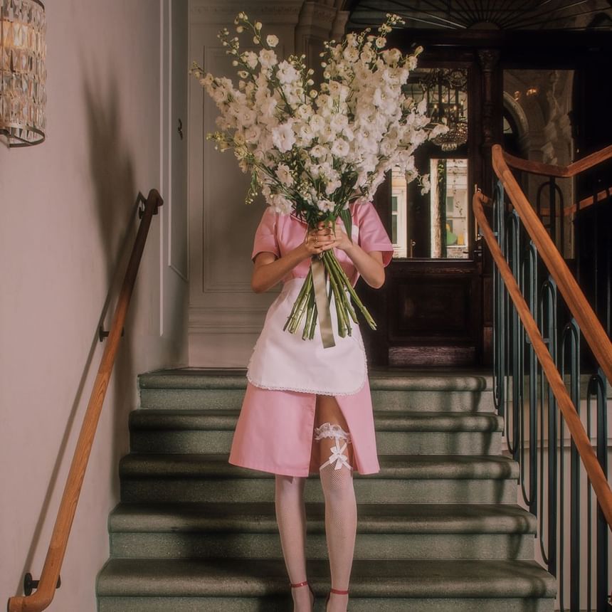 Woman in a pink maid dress holding a large bouquet of white flowers, standing on a staircase at Hotel Motto Vienna