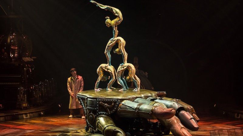Cirque du Soleil artists perform acrobatics on a giant mechanical hand in steampunk style.
