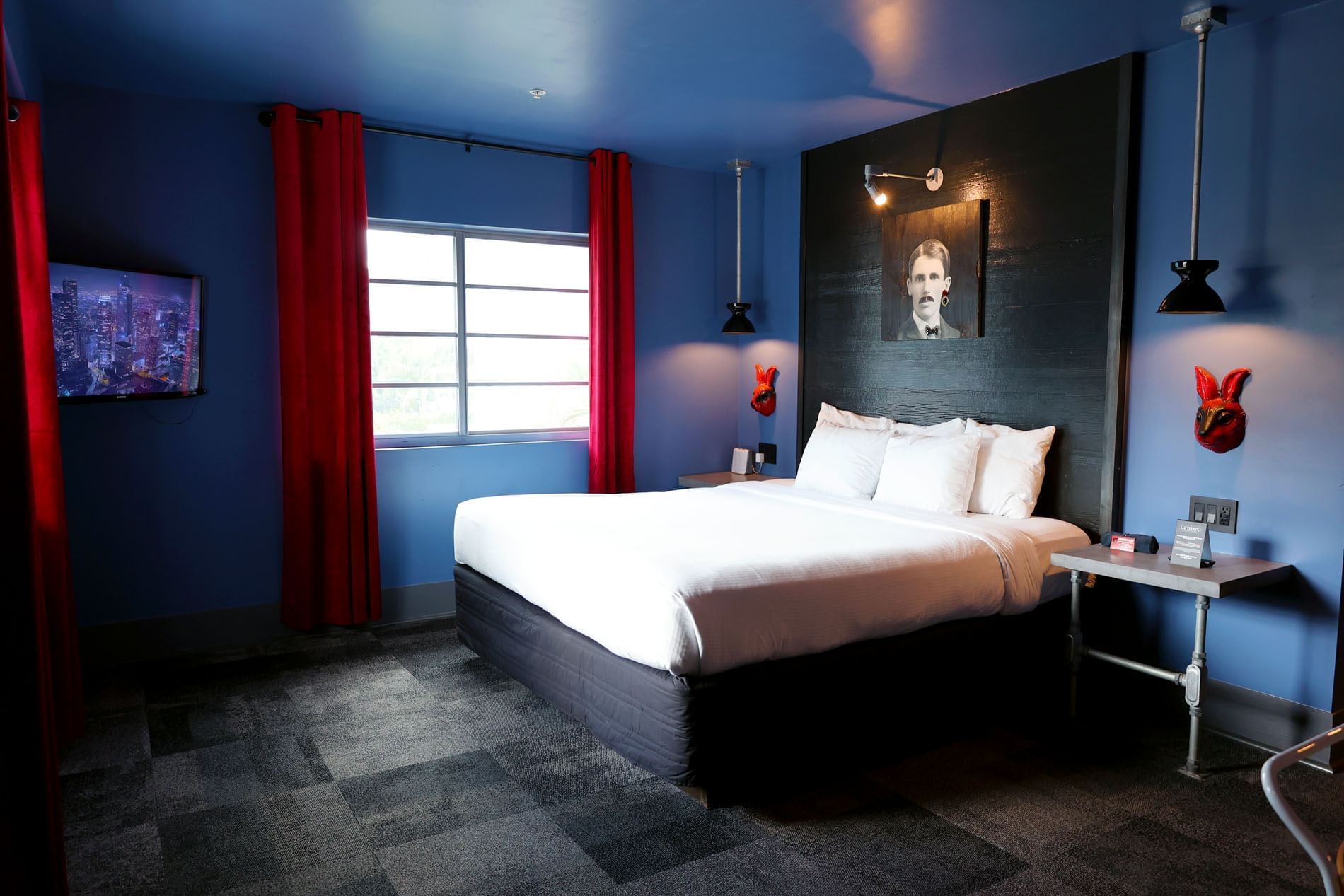 A hotel room with a king-sized bed, blue walls, red curtains, and a flat-screen TV.