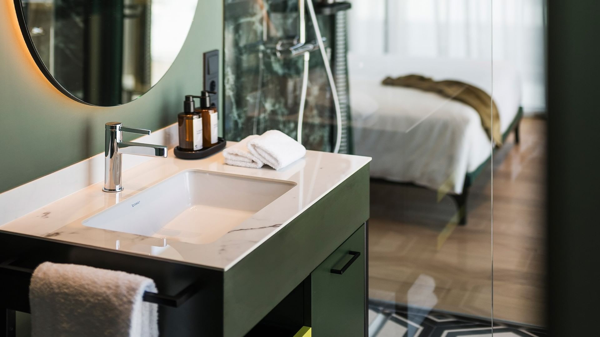 Bagno con lavandino e letto sfuocato in Camera Grand Deluxe al Falkensteiner Hotel Bozen WaltherPark di Bolzano.