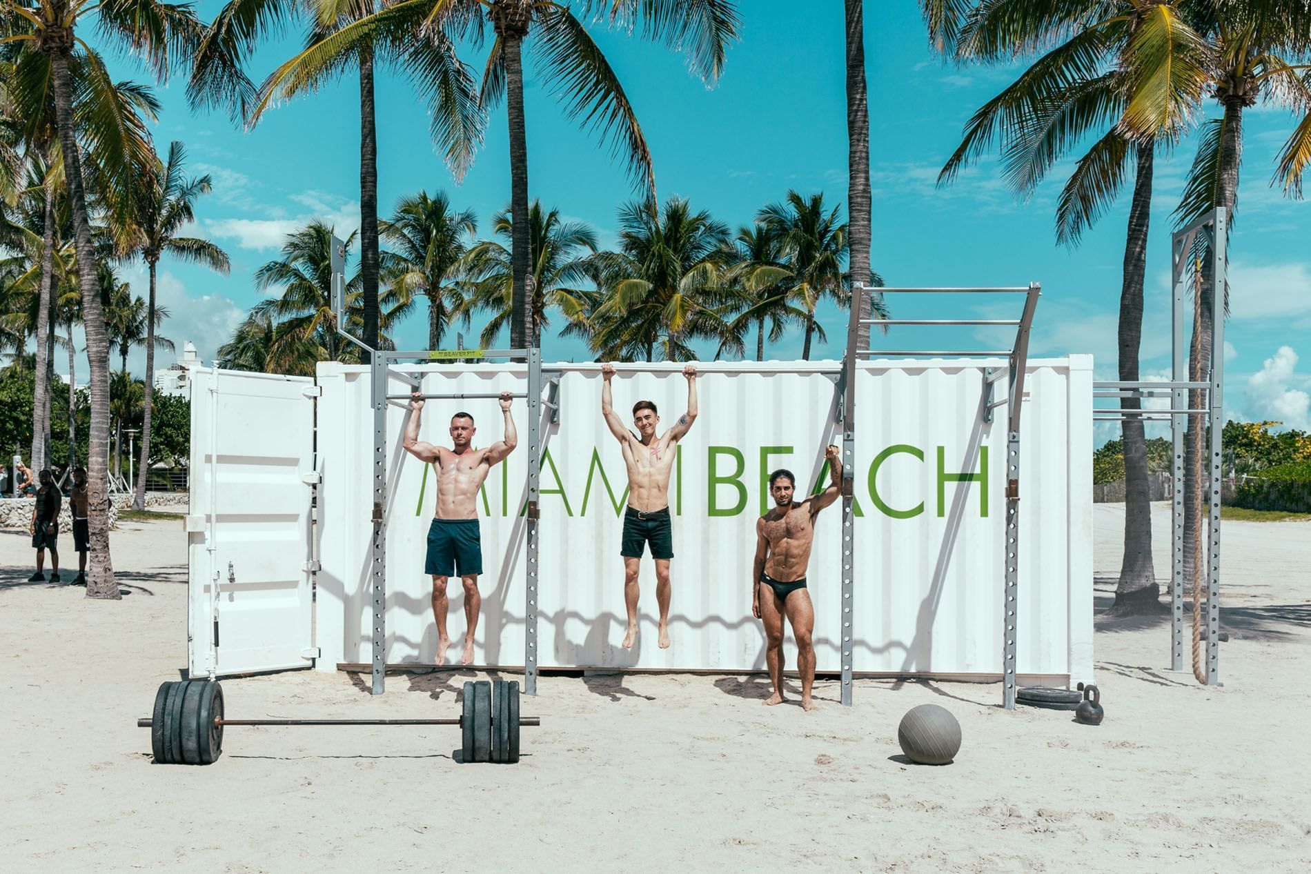 Three men exercising on a beach in front of a white shipping container with 'MIAMI BEACH' written on it.